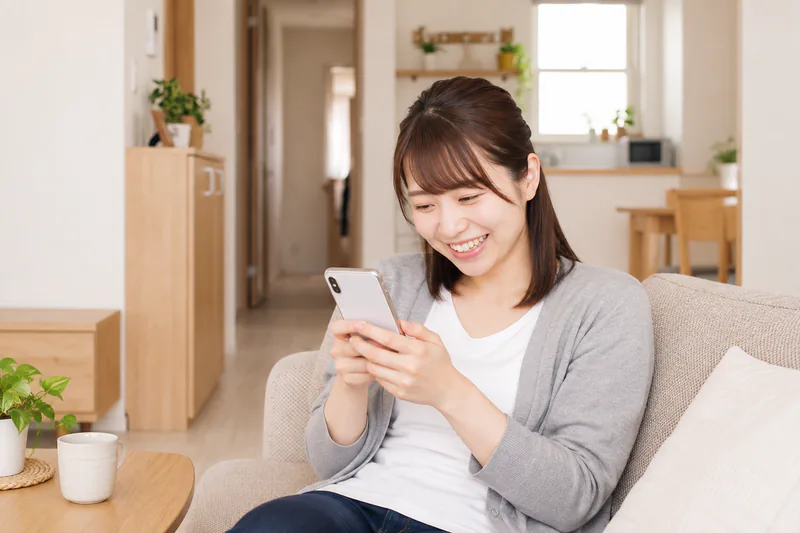 ソファでスマートフォンを見て微笑む女性（イメージ）